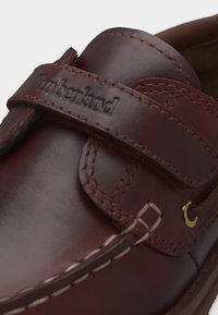 Κοντινή λήψη καφέ δερμάτινου παπουτσιού Timberland με ραμμένες λεπτομέρειες και ανάγλυφο λογότυπο στο λουράκι επάνω.