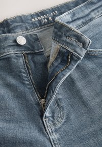 Siniste teksapükstega, millel on barrel-lõige, lukuga eesmine osa, metallist nööp ja kontrastsed õmblused. Sile tekstuur ja keskmine pesutöötlus.