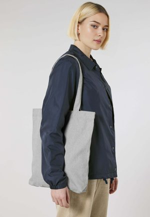 Borsa tote in tessuto grigio con un design semplice, di grande dimensione e manici lunghi, indossata sopra una giacca blu navy, creando un look informale.