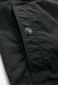 Tissu noir avec un gros plan d'une poche à rabat cousue maintenue par un bouton en métal rond portant l'inscription "Produits Durables de Qualité Supérieure".