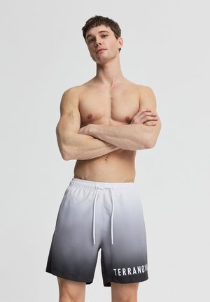 Giovane uomo senza maglietta con le braccia incrociate che indossa pantaloncini sfumati dal bianco al nero con coulisse bianca e scritta "TERRANOVA" sulla gamba sinistra.