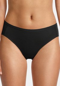 Donna che indossa slip in cotone neri a vita media, mostrando il torso e le cosce superiori su uno sfondo bianco.