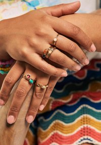 Goldringe mit bunten Edelsteinen in Rot, Grün und Schwarz. Ein Ring hat einen Perlenakzent. Hände liegen auf einer mehrfarbigen, gemusterten Oberfläche.