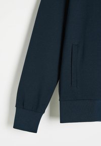 Granatowy sweter z gładką fakturą, ściągaczami przy mankietach i małą kieszonką z boku. Design jest minimalistyczny i dopasowany.