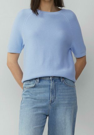 Vrouw draagt een lichtblauwe, kortmouwige gebreide trui, ingestopt in een high-waisted lichtblauwe spijkerbroek, met haar handen achter haar rug.