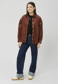 Chemise en velours côtelé marron avec de gros boutons, portée sur un pull noir, associée à un jean denim foncé et des baskets crème avec des bordures grises.