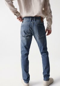 Chinos de mezclilla azul con un corte ajustado, bolsillos traseros y un ligero desvanecimiento. Combinados con una camisa beige de manga larga y zapatillas blancas.
