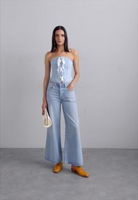 Denim top brez naramnic z belim trakcem, v kombinaciji z ohlapnimi svetlo modrimi kavbojkami in rumeno-oranžnimi visokimi čevlji. Drži belo torbico.