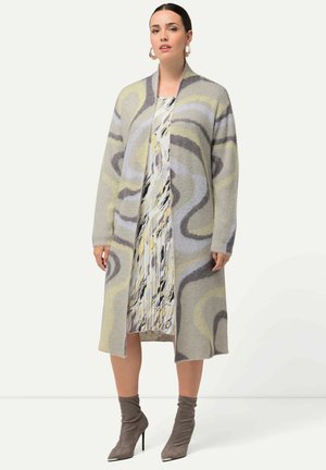 Langer Cardigan in sanftem Grau mit abstrakten Wirbelmustern in Gelb und Braun. Kombiniert mit einem plissierten, gemusterten Kleid und grauen Knöchelstiefeln.