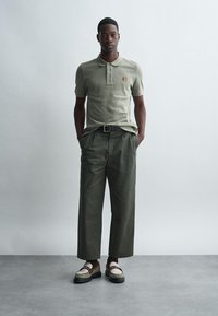 Man standing with hands in pockets wearing light olive polo, dark olive wide-leg trousers, and two-tone loafers on gray floor.