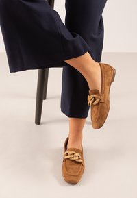 Mocasines de gamuza marrón con detalle de tira trenzada texturizada. Material suave y forma de punta redondeada, combinados con pantalones de pierna ancha en color azul marino.
