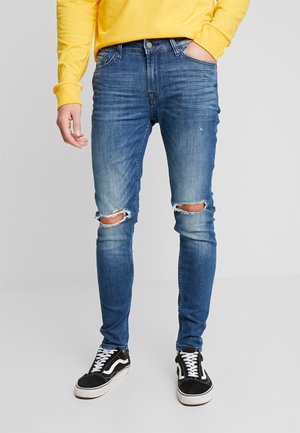 Person trägt blaue zerrissene Skinny-Jeans, schwarz-weiße Turnschuhe und ein gelbes Sweatshirt, eine Hand in der Tasche.
