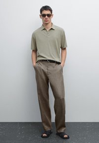Polo shirt verde oliva chiaro con maniche corte, pantaloni in lino marrone e sandali neri a scivolo. Il modello indossa occhiali da sole rettangolari.