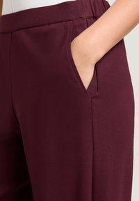 Pantalones burdeos hechos de tejido elástico, con un bolsillo lateral y una cinturilla elástica para mayor comodidad. Diseño suave y limpio sin patrones visibles.