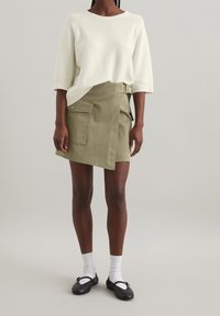 Pull en maille beige clair avec des manches larges associé à une jupe portefeuille vert olive dotée d'une poche latérale et d'un tissu texturé.