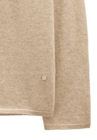 Beżowy sweter dzianinowy o miękkiej teksturze, z małą metalową metką z logo marki w pobliżu dolnej krawędzi oraz ściągaczami na rękawach i dolnej krawędzi.