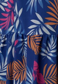 Niebieska tkanina z tropikalnym wzorem liści w odcieniach różu, pomarańczu i bieli. Posiada falbaniasty szew oraz gładką teksturę.