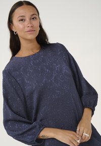 Marinblus med texturerad tyg, rund halsringning och långa puffärmar. Har subtila mönster och elastiska ärmslut.