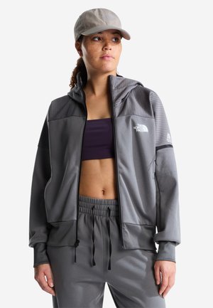 Kobieta ubrana w szarą kurtkę North Face na suwak, dopasowane szare spodnie, ciemny sportowy biustonosz i beżową czapkę z daszkiem, patrząca na bok.