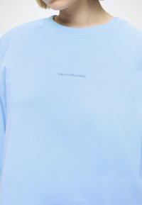 Personne portant un sweat-shirt bleu clair avec un petit texte "Marc O'Polo DENIM" brodé sur la poitrine, debout devant un fond uni.