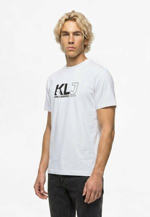 Blond muž s krátkymi vlnitými vlasmi, oblečený v biele tričko Karl Lagerfeld Jeans a tmavých džínsoch, stojaci pred jednoliatym bielym pozadím.