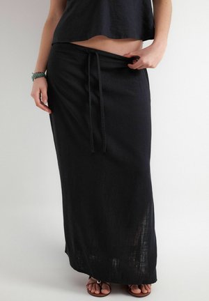 Žena si upravuje pás dlhej čiernej sukne so šnúrkou, má na sebe čierny krátky top a sandále, na jednoliatom bielom pozadí.