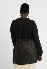 Sort gennemsigtig bluse med tekstureret stof og små ophøjede prikker, lange ærmer og en samlet talje. Elegant, let design.