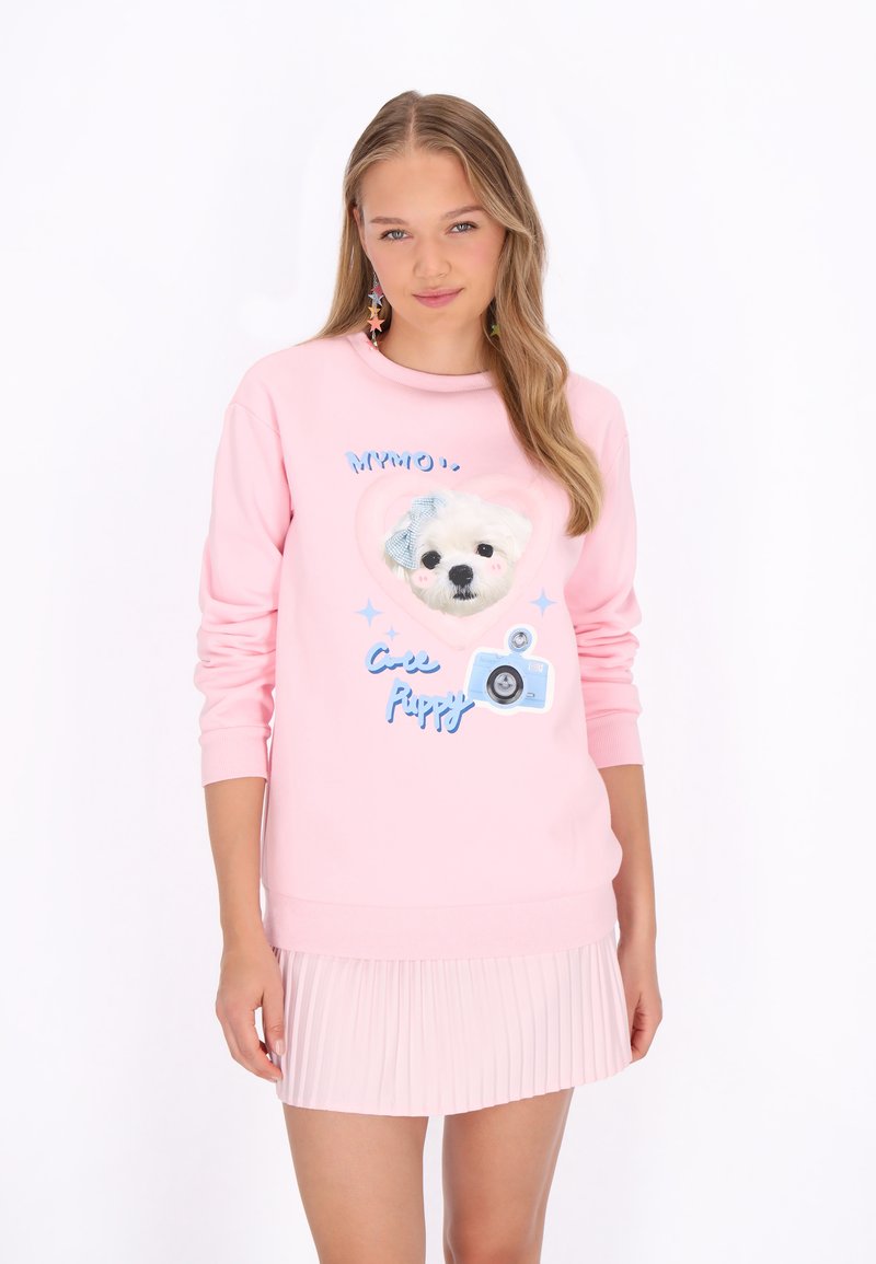 Világos rózsaszín pulóver, amely egy kölyökkutya arcát és egy fényképezőgépet ábrázoló grafikával, kék díszítésekkel és hosszú ujjakkal rendelkezik. Alul pliszírozott szoknya látható.