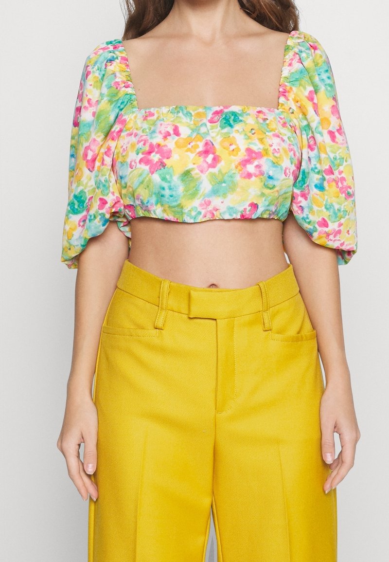 Femme portant un crop top à manches bouffantes fleuries avec un pantalon jaune taille haute, debout devant un fond uni.