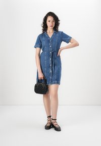 Denim srajčna obleka s kratkimi rokavi, gumbi spredaj in pasom. Črna teksturirana torbica z dvema ročajema, nošena na desni strani.