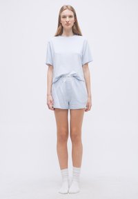 Νεαρή γυναίκα σε όρθια στάση, φορώντας ανοιχτό μπλε κοντομάνικο τοπ και σορτς που ταιριάζουν, με λευκές κάλτσες σε απλό φόντο.