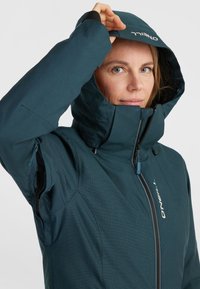 Teal waterproof jacket with a high collar and adjustable hood. Features a front zipper, side pockets, and textured fabric. Logo at center.