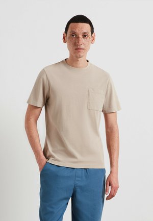 Beige katoenen T-shirt met korte mouwen en een borstzakje, gecombineerd met lichtblauwe broek. Soepele textuur, klassieke ronde hals.