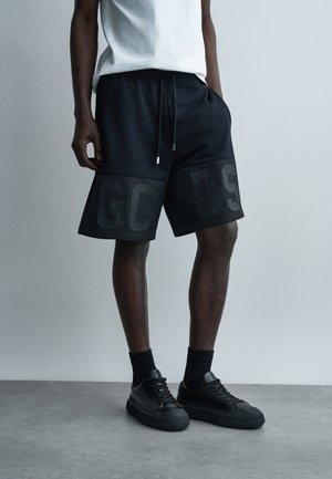 Άνδρας που φοράει μαύρα σορτς GCOS με κορδόνι, λευκό t-shirt, μαύρες κάλτσες και μαύρα αθλητικά παπούτσια, στέκεται σε γκρι δάπεδο μπροστά από απλό τοίχο.