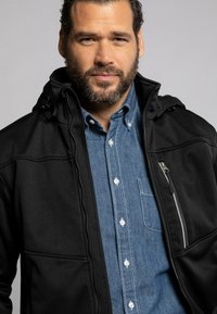 Schwarze Softshelljacke mit einem Frontreißverschluss, verstellbarem Kapuzen und einer Reißverschlusstasche. Über einem hellblauen Denimhemd mit Knopfleiste getragen.