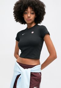 Čierne skrátené tričko s okrúhlym výstrihom, krátkymi rukávmi a malým logom Nike; nosené s marhuľovými šortkami a svetlomodrou mikinou viazanou v pase.