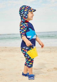 Costume da bagno per bambini con modelli di squali blu e multicolori, accenti blu, secchiello coordinato e scarpe blu, posati su una spiaggia di sabbia.