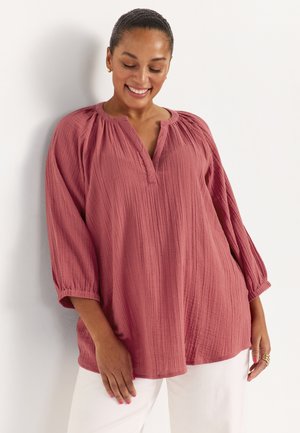 Lachende vrouw met donker haar in een knot, gekleed in een loszittende, gestructureerde blouse in stofroze en witte broek tegen een witte achtergrond.