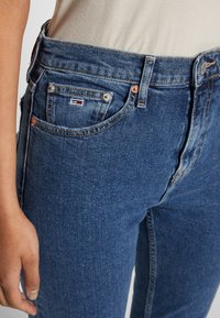 Dark blue denim jeans feature a classic five-pocket design, copper-tone hardware, and a small logo label on the front pocket.