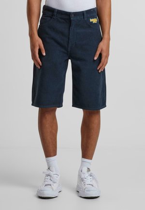 Dunkelblaues Cordshorts mit geradem Schnitt und elastischem Bund, mit einem gelben gestickten Logo auf der Vorderseite. Weiße Sneaker und Socken runden den Look ab.
