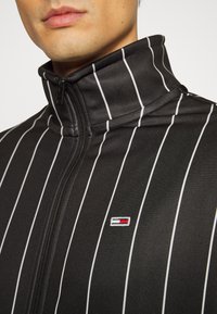 Svart zip-up jacka med vita vertikala ränder, hög krage och broderad logotyp i rött, vitt och blått på vänster sida. Slät textur.
