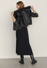 Donna con giacca in pelle trapuntata nera, gonna lunga nera e stivali neri, che sistema il colletto della giacca mentre si trova in piedi contro uno sfondo chiaro.