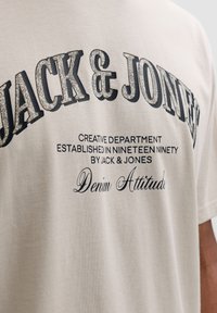 Ανοιχτό μπεζ βαμβακερό T-shirt με έντονα μαύρα γράμματα "JACK & JONES" στο μπροστινό μέρος, καθώς και μικρότερες λεπτομέρειες κειμένου από κάτω.