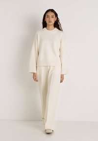 Krém színű kötött pulóver bordázott szegéllyel, hozzá illő, bő szárú nadrággal párosítva. Puha tapintású, laza szabású, egyszerű sziluett.
