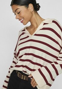 Maronen- und crèmefarbener gestreifter Pullover aus Strick mit V-Ausschnitt und weiten Ärmeln, kombiniert mit einem braunen Gürtel und schwarzen Hosen. Pflanzen im Hintergrund sichtbar.