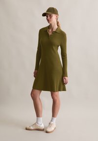 Abito lungo a maniche lunghe verde oliva a coste, con colletto e bottoni. Abbinato a calzini bianchi e sneakers beige, con un cappellino abbinato.