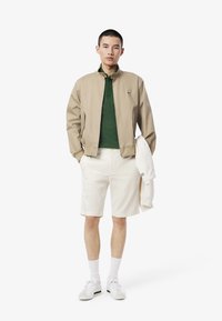 Giubbotto bomber beige sopra una polo verde, abbinato a pantaloni corti bianchi. Sneakers bianche con dettagli verdi. Design minimalista, texture morbida.