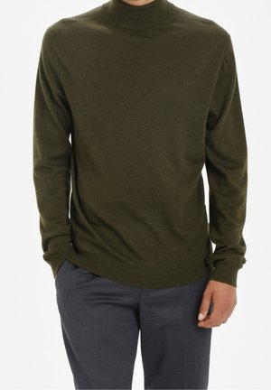 Mężczyzna w oliwkowozielonym golfie z długimi rękawami i ciemnoszarych spodniach, ręce swobodnie opuszczone wzdłuż ciała.