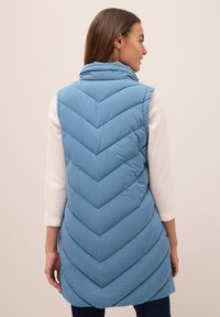 Femme portant un gilet sans manches matelassé bleu clair avec un motif en chevrons, un haut à manches longues crème et un pantalon foncé, de dos et souriante.