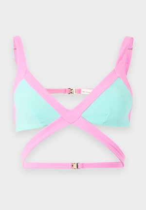 Kvinde iført blå og pink todelt badedragt med udskæringer, iført hvide sandaler med høje hæle mod en ensfarvet hvid baggrund.